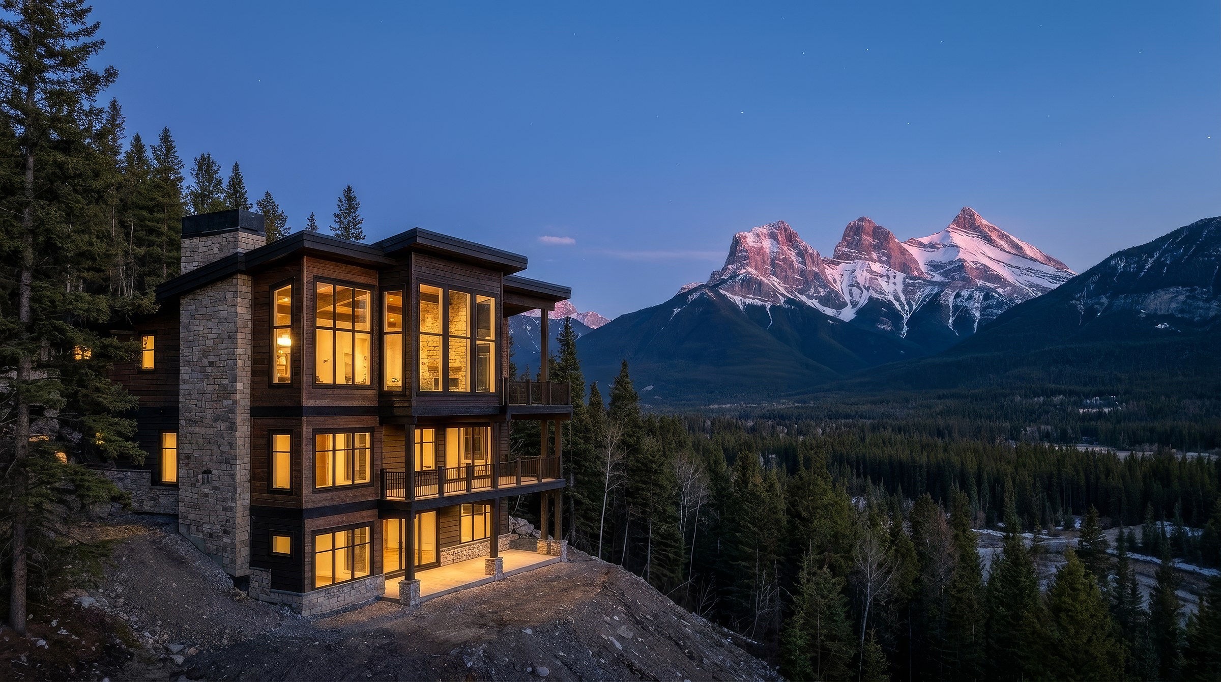 Serenity Point at dusk in Silvertip, Canmore, its windows lit, with the Three Sisters across the Bow Valley.