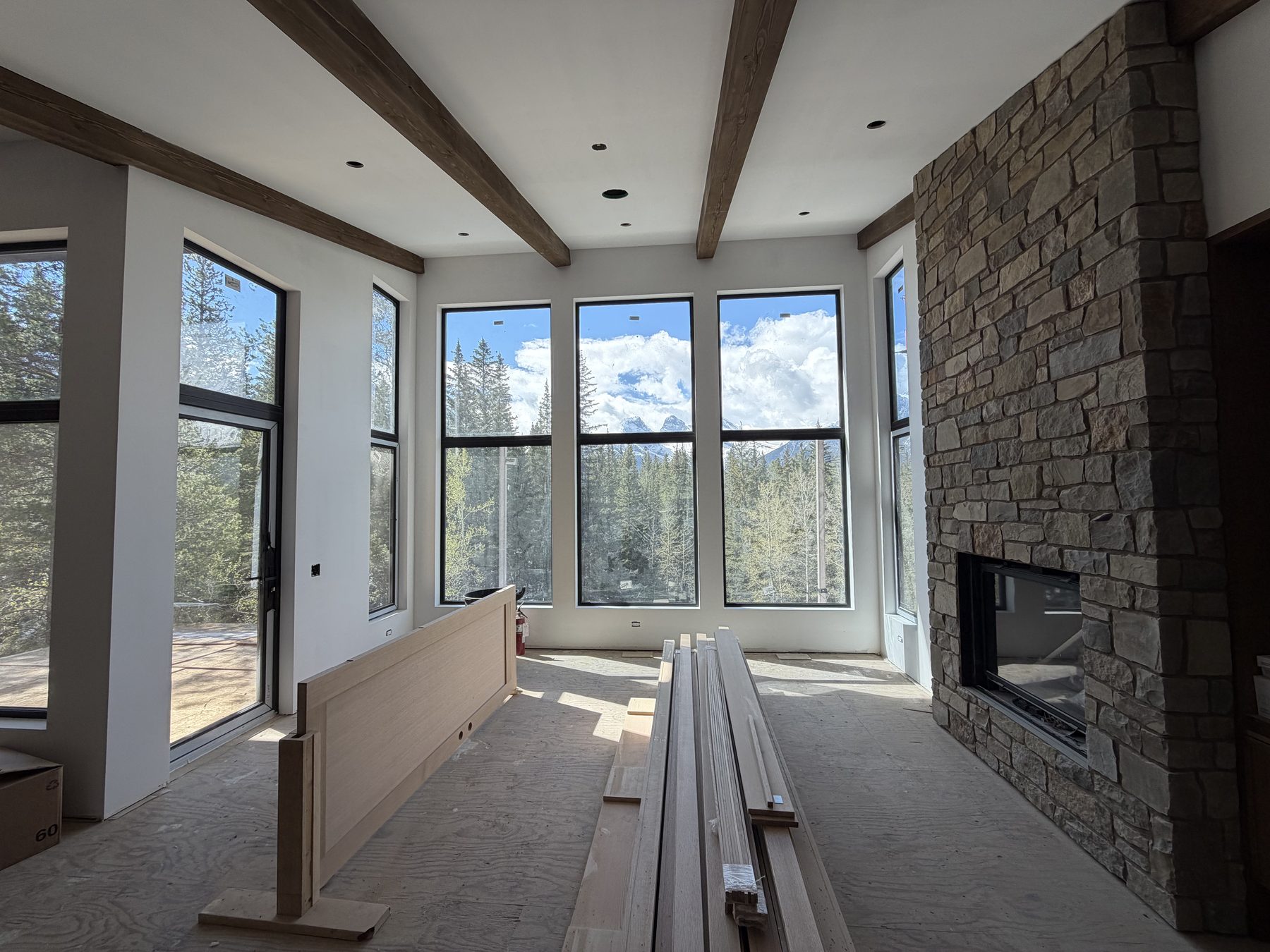 Great room under construction at Serenity Point. Floor-to-ceiling south-facing windows framing the mountains and a stone fireplace on the right wall.